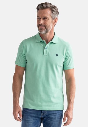 Skægget mand iført en mintgrøn poloshirt med to knapper og et lille navyblåt logo, kombineret med blå jeans, stående mod en hvid baggrund.