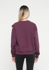 Sweatshirt violet à coupe décontractée, avec des accents de volants aux épaules et un ourlet et des poignets côtelés, porté sur des leggings noirs.