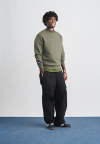 Alyvuogių žalia džemperis su apvalia iškirpte, dėvimas ant žalsvos spalvos marškinių; derinamas su laisvais juodais karinėmis kelnėmis ir juodais sportbačiais. Matoma tekstūruota medžiaga.