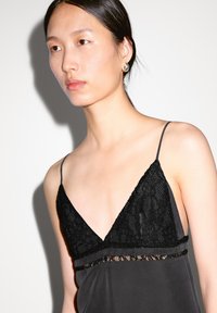 Black satin camisole featuring a deep V-neck with lace trim, thin straps, and a delicate lace accent at the waist. Smooth texture.