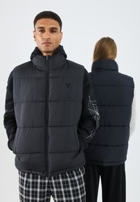 Gilet rembourré noir avec col haut, fermeture éclair et poches avant. Présente des manches contrastantes avec un motif de toile d'araignée blanche.