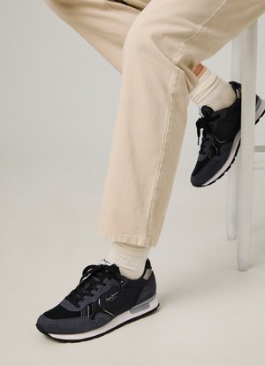Zwarte en grijze sneakers met een mix van stof en suede, voorzien van veters, een gewatteerde kraag en een witte tussenzool, gedragen met beige broek en witte sokken.