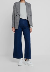 Blazer à carreaux gris à double boutonnage sur un haut gris clair, associé à un jean bleu foncé à jambes larges et des baskets blanches.