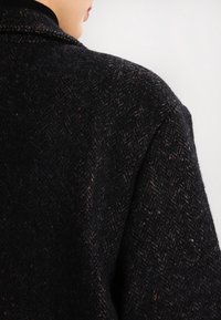 Černý kabát s herringbone vzorem vyrobený z texturované vlněné tkaniny. Má široké ramena a klasický límec.