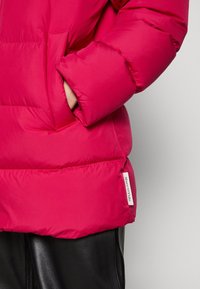 Chaqueta acolchada fucsia con textura suave, que presenta un bolsillo lateral y una etiqueta de marca en el dobladillo. La manga tiene un detalle de puño fruncido.