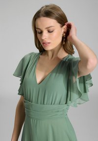 Groene chiffon jurk met fluttermouwen, diepe V-hals en een getailleerde taille. De stof is glad met een vloeiend ontwerp en een lichte textuur.
