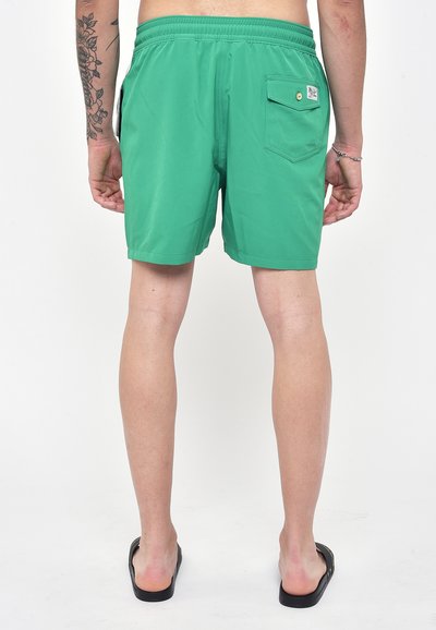 Pantaloni da nuoto verdi con vita elastica, tasca posteriore e texture liscia. Indossati con sandali neri a slip.