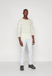 Man standing facing forward wearing a cream long-sleeve shirt, light gray pants, and dark gray sneakers against a plain white background.