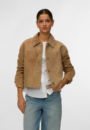 Femeie cu păr închis la culoare purtând o jachetă maro deschis din piele întoarsă peste o cămașă albă cu nasturi și blugi albaștri deschiși, stând în picioare împotriva unui fundal simplu.