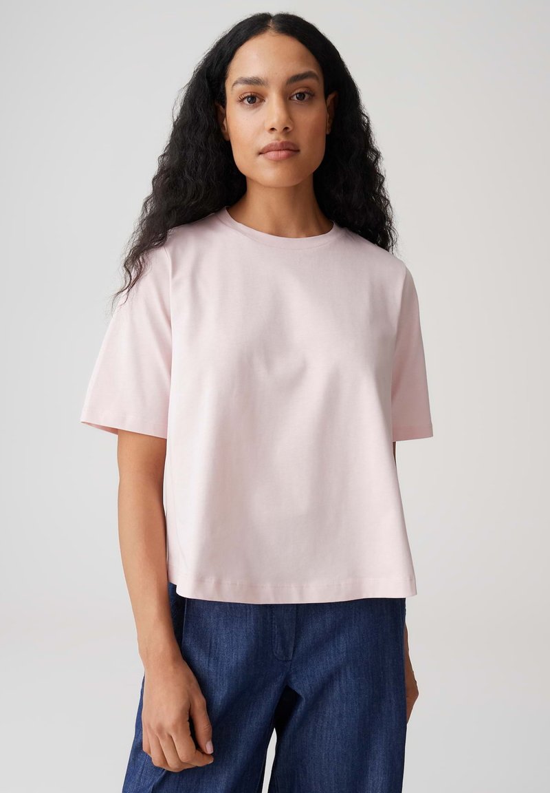 Femme aux cheveux longs et bouclés portant un t-shirt rose pâle à manches courtes et un jean bleu foncé, debout devant un fond clair uni.