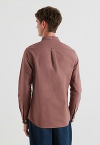 Een blouse met lange mouwen van stof in stoffige rozenkleur, met een knoopsluiting kraag, een rugplooien en knoopmanchetten. Glad oppervlak en op maat gemaakte uitstraling.