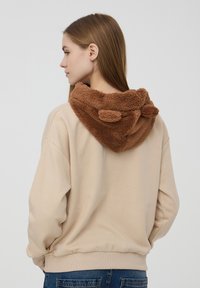 Felpa di pile beige con cappuccio marrone e morbido. Il cappuccio è realizzato in un tessuto testurizzato e presenta dettagli arrotondati nella parte superiore. Vestibilità comoda e polsini elastici.