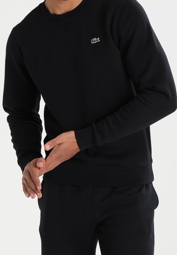 Sudadera negra de manga larga, cuello redondo, puños y cintura acanalados. Presenta un pequeño logo verde de Lacoste en el lado izquierdo del pecho.