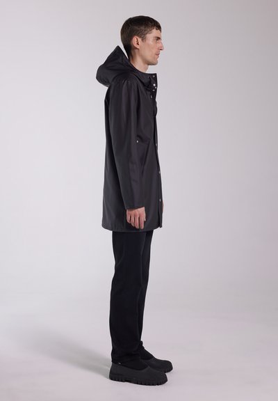 Black waterproof jacket with a hood, front snap closure, and two side pockets. Worn over black pants and chunky black shoes. Side view.