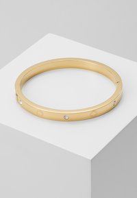Bracelet jonc en finition dorée, avec une surface lisse, présentant trois cristaux transparents incrustés et des accents de logo gravés. Design rond, ouvert.