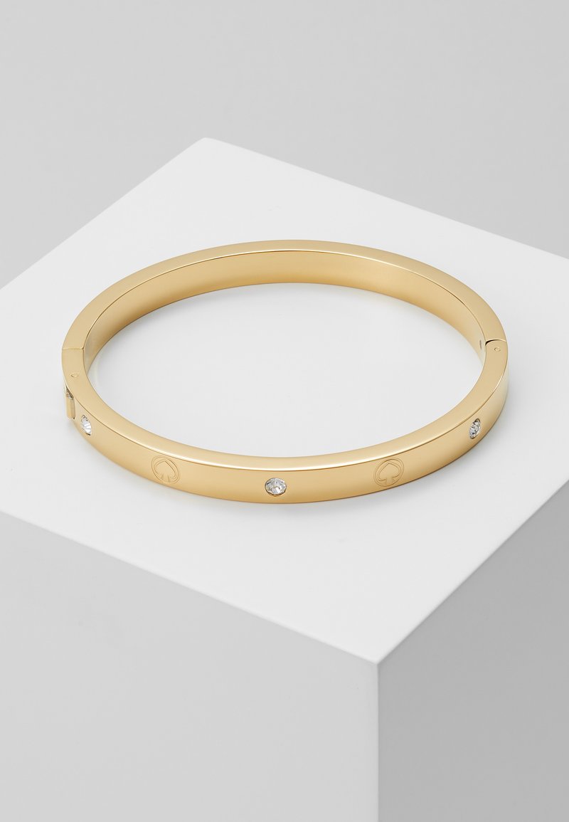 Bracelet jonc en finition dorée, avec une surface lisse, présentant trois cristaux transparents incrustés et des accents de logo gravés. Design rond, ouvert.