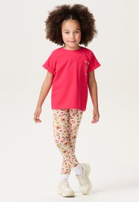 T-shirt en coton rouge avec des manches retroussées, associée à des leggings roses à motif floral. L'enfant porte des baskets blanches avec des semelles rembourrées.