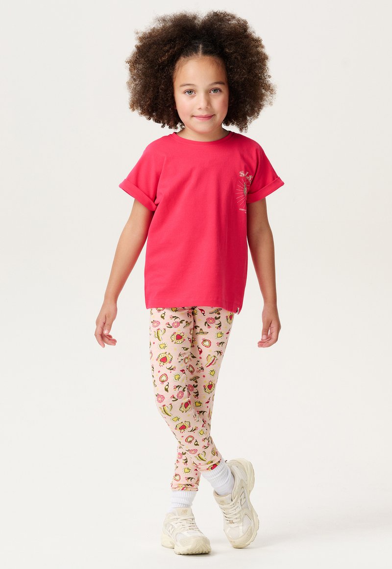 T-shirt en coton rouge avec des manches retroussées, associée à des leggings roses à motif floral. L'enfant porte des baskets blanches avec des semelles rembourrées.