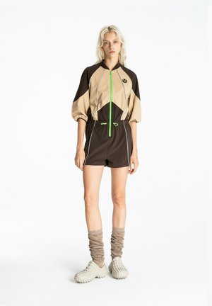 Mujer joven con un mono beige y marrón con cremallera y detalles verdes, calcetines beige arrugados y zapatos beige gruesos, de pie sobre fondo blanco.