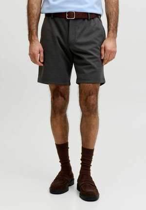 Mann trägt dunkelgraue Shorts, braunen Gürtel, hellblaues Hemd, braune Socken und braune Wildleder-Loafer, steht vor einem einfarbigen Hintergrund.