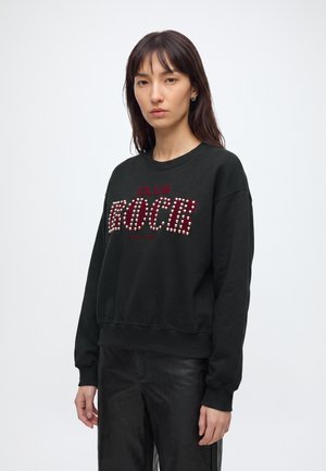 Femme portant un sweat-shirt noir avec "CLUB ROCCS" en clous rouges et blancs, associé à un pantalon noir, debout devant un fond blanc uni.