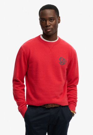 Mand iført en rød sweatshirt med et lille sort logo på brystet, mørke bukser, brunt flettet bælte og et armbåndsur, stående mod en hvid baggrund.