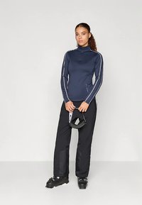 Donna che indossa un top a maniche lunghe blu navy e pantaloni da neve neri, tiene in mano degli occhiali da sci neri, in piedi su uno sfondo bianco uniforme.