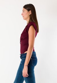 Body aderente color bordeaux con maniche a cappuccio, design sagomato. Abbinato a jeans blu scuro, che mostrano una texture liscia e una silhouette snella.