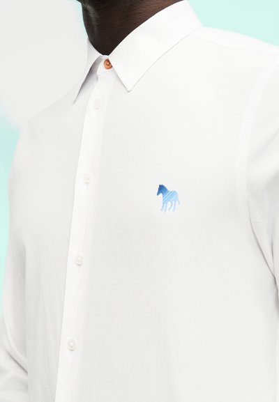 Άνδρας που φοράει λευκό πουκάμισο με κουμπιά και έχει κεντημένο μπλε λογότυπο αλόγου στο αριστερό στήθος, σε φωτεινό φόντο.