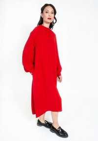 Červené šaty s dlouhým rukávem, vysokým límcem, uvolněným střihem a nařasenými detaily, doplněné černými lesklými nazouvacími botami. Hladká textura.