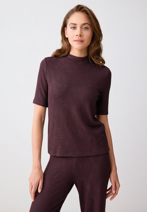 Donna che indossa una maglietta a maniche corte in tessuto strutturato di colore viola scuro e pantaloni abbinati, in piedi davanti a uno sfondo semplice di colore grigio chiaro.