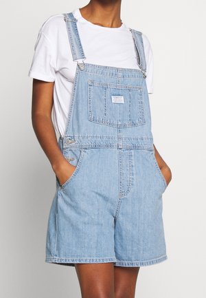 Person iført lyseblå denim korte overaller og en hvid t-shirt med korte ærmer med hænderne i lommerne mod en ensfarvet baggrund.