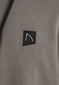 Grijze hoodie met een gladde textuur, voorzien van een klein zwart patch met een wit geometrisch lijndesign en een zwarte koord.