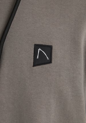 Felpa grigia con una texture liscia, caratterizzata da un piccolo patch nero con un design geometrico bianco e un laccio nero.