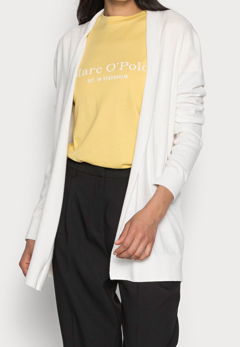 Personne portant un t-shirt jaune pâle Marc O’Polo, un cardigan blanc ouvert et un pantalon noir taille haute devant un fond uni.