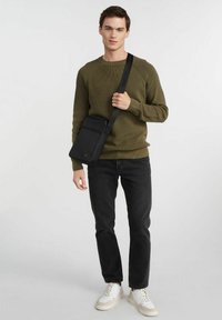 Czarna torba na ramię z regulowanym paskiem, z przednią kieszenią, eleganckim designem i fakturowaną powierzchnią, noszona przez osobę w oliwkowym swetrze.