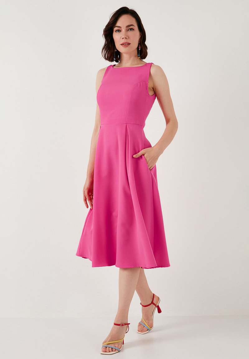 Rochie roz ajustată, cu decolteu pătrat, design fără mâneci, buzunare laterale și fustă evazată. Modelul poartă sandale cu curelă multicolore.