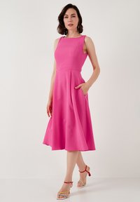 Fitted pink dress with a square neckline, sleeveless design, side pockets, and a flared skirt. Model wears multicolored strappy sandals.
