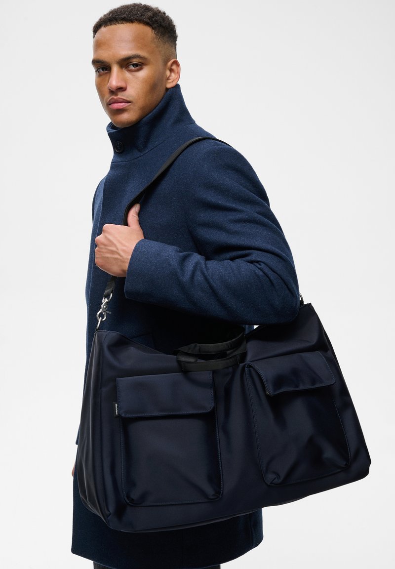 Homme portant un manteau bleu foncé et une grande sacoche bleu marine avec deux poches frontales sur l'épaule, sur fond blanc.