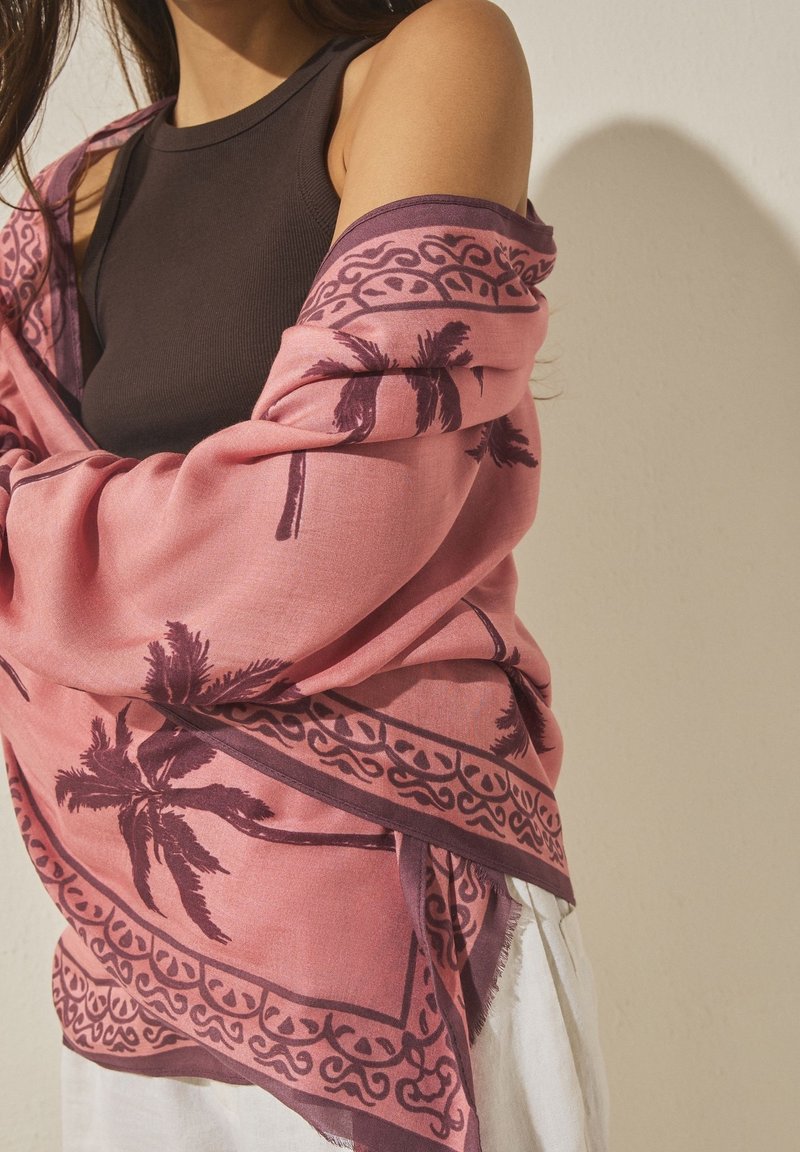 Femme portant un top noir sans manches et un châle rose avec des motifs sombres de palmiers et décoratifs drapé sur ses épaules.