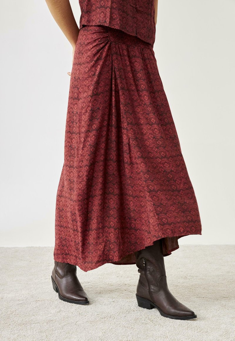 Falda larga de patrón floral rojo, hecha de una tela ligera, combinada con botines de cuero marrón que presentan detalles texturizados y tacones apilados.