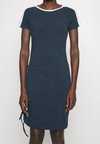 Tummanlaivansiniset lyhythihaiset mekko, jossa on pyöreä kaula-aukko ja valkoiset yksityiskohdat olkapäillä. Mekossa on sivuhauska helmassa säätömahdollisuus.