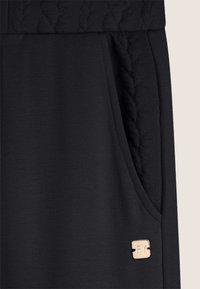 Pantalones deportivos negros con una cinturilla texturizada y un bolsillo lateral, y una etiqueta de logo en bronce en el borde inferior. Tejido suave con un acabado mate.