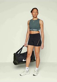 Γυναίκα σε αθλητική ενδυμασία, κρατώντας μια μαύρη τσάντα duffel της Nike, στέκεται με τα μάτια κλειστά σε έναν απλό ανοιχτό φόντο.