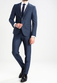 Granatowy garnitur z subtelnymi wzorami, jednorzędową marynarką, klapami z wycięciem w szpic i dopasowanym krojem, zestawiony z białą koszulą i ciemnym krawatem.