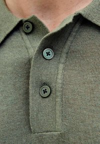 Olive zaļas krāsas adīta polo krekls ar trim pogu aizdari; ar apaļām pogām tumši zaļā krāsā ar melnām caurumiem un faktūru audumu.