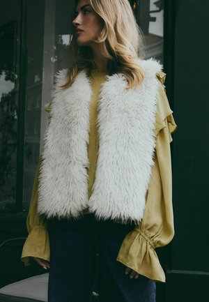 Hvid kunstpels vest over en sennepsgul bluse med flæseærmer, parret med mørkeblå denim. Baggrunden har en grøn væg.