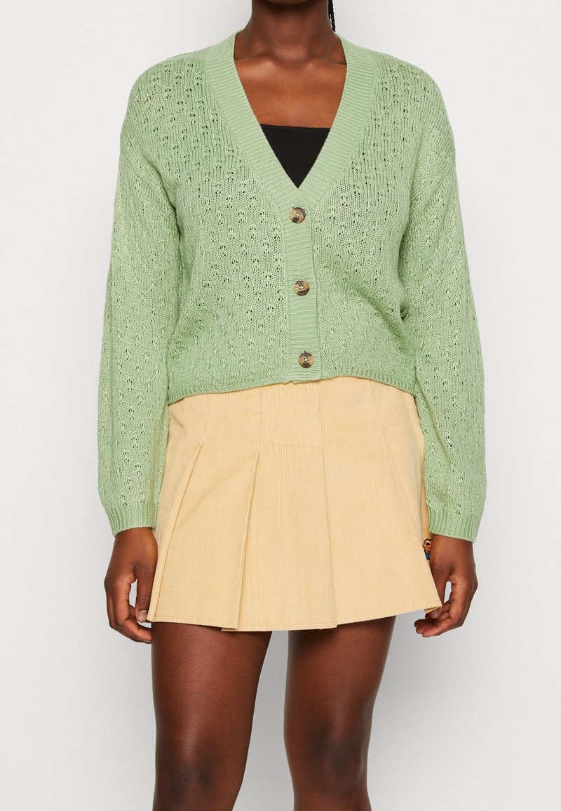 Personne portant un cardigan en maille vert clair boutonné par-dessus un haut noir et une mini-jupe plissée beige, debout devant un fond uni.