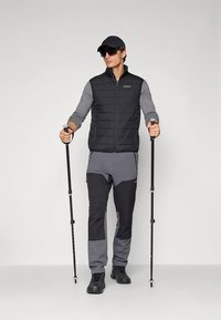 Schwarze wattierte Weste über einem grauen Langarmshirt, kombiniert mit grauen und schwarzen Hosen. Die Person hält zwei Trekkingstöcke und trägt schwarze Schuhe.