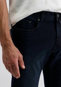Männliche Hand entspannt neben dunkelblauen Jeans und einem weißen Hemd, sichtbar sind die Fronttasche und der Taillenbereich.
